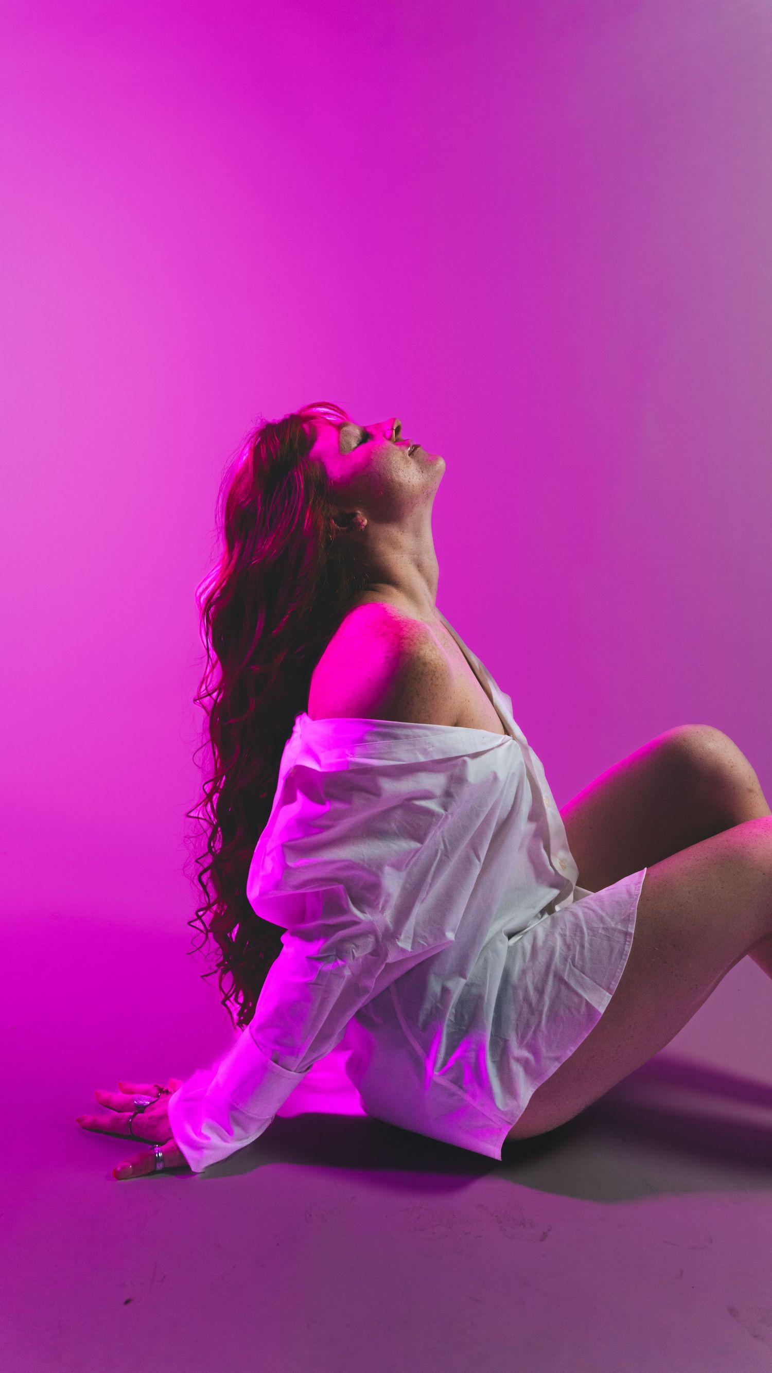 Woman with long curly hair posing on floor against vibrant pink background in white outfit.
