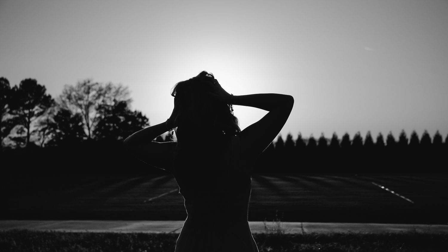 Silhouette of woman with hands in hair against sunset sky in black and white photo.