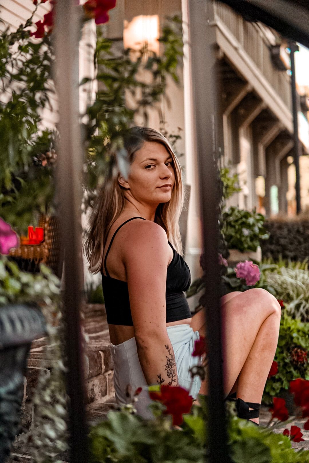 Blonde woman in black crop top sitting among colorful garden flowers on urban porch steps.
