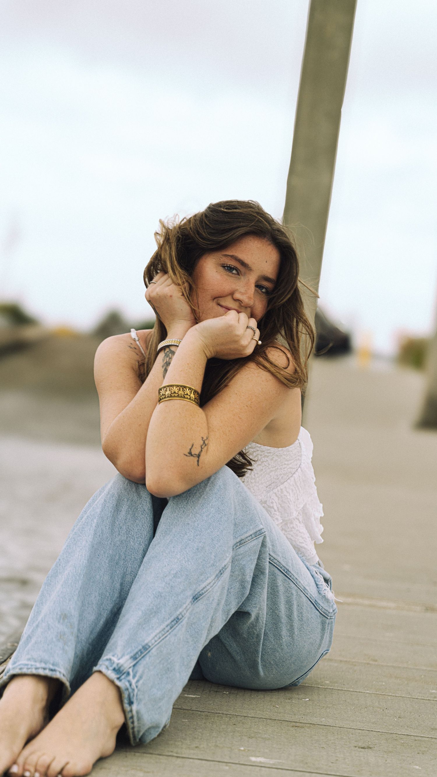 Young woman with brown hair sitting on wooden boardwalk, smiling, wearing white top and light blue jeans.