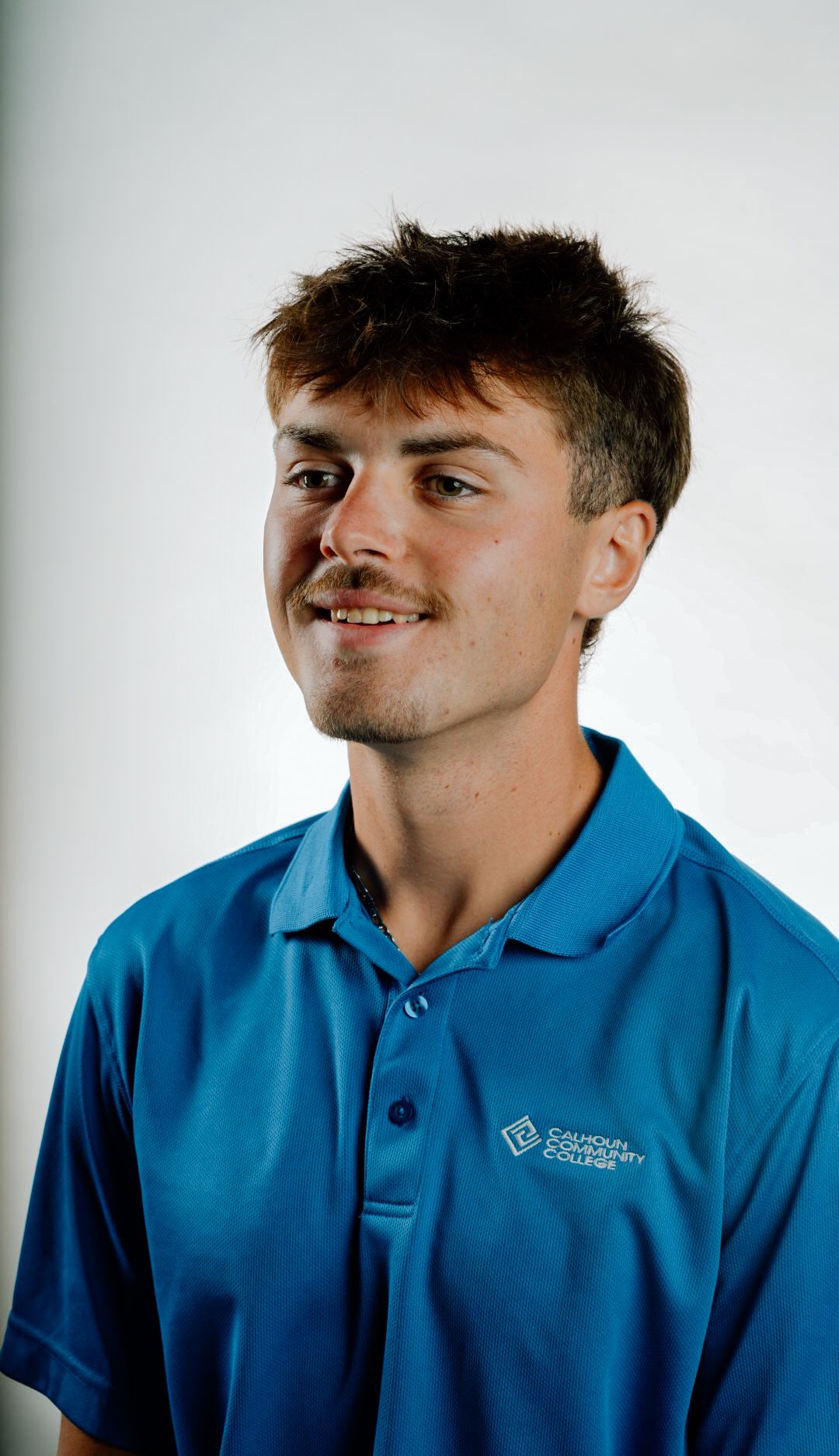 Smiling young man with dark hair wearing blue Calhoun Community College polo shirt against white background.