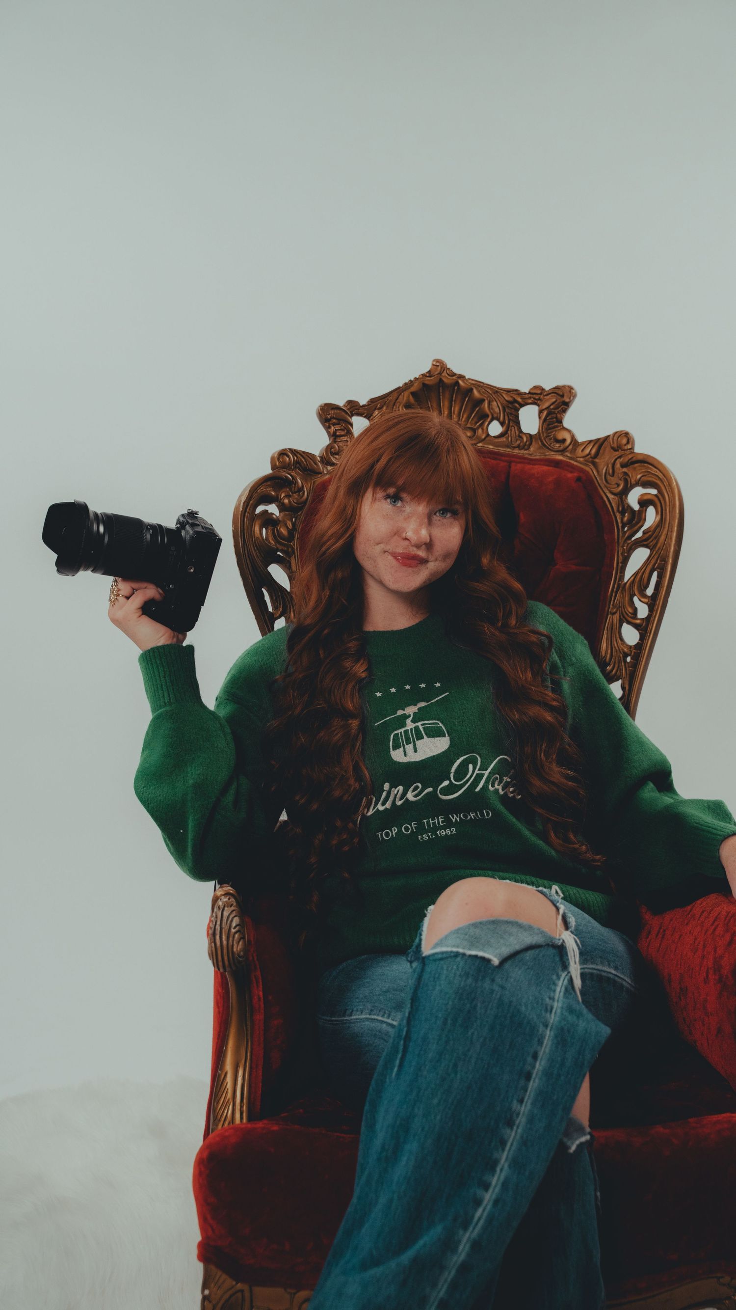 Young woman with red hair in green sweatshirt holding camera, seated in ornate throne chair, wearing ripped jeans.