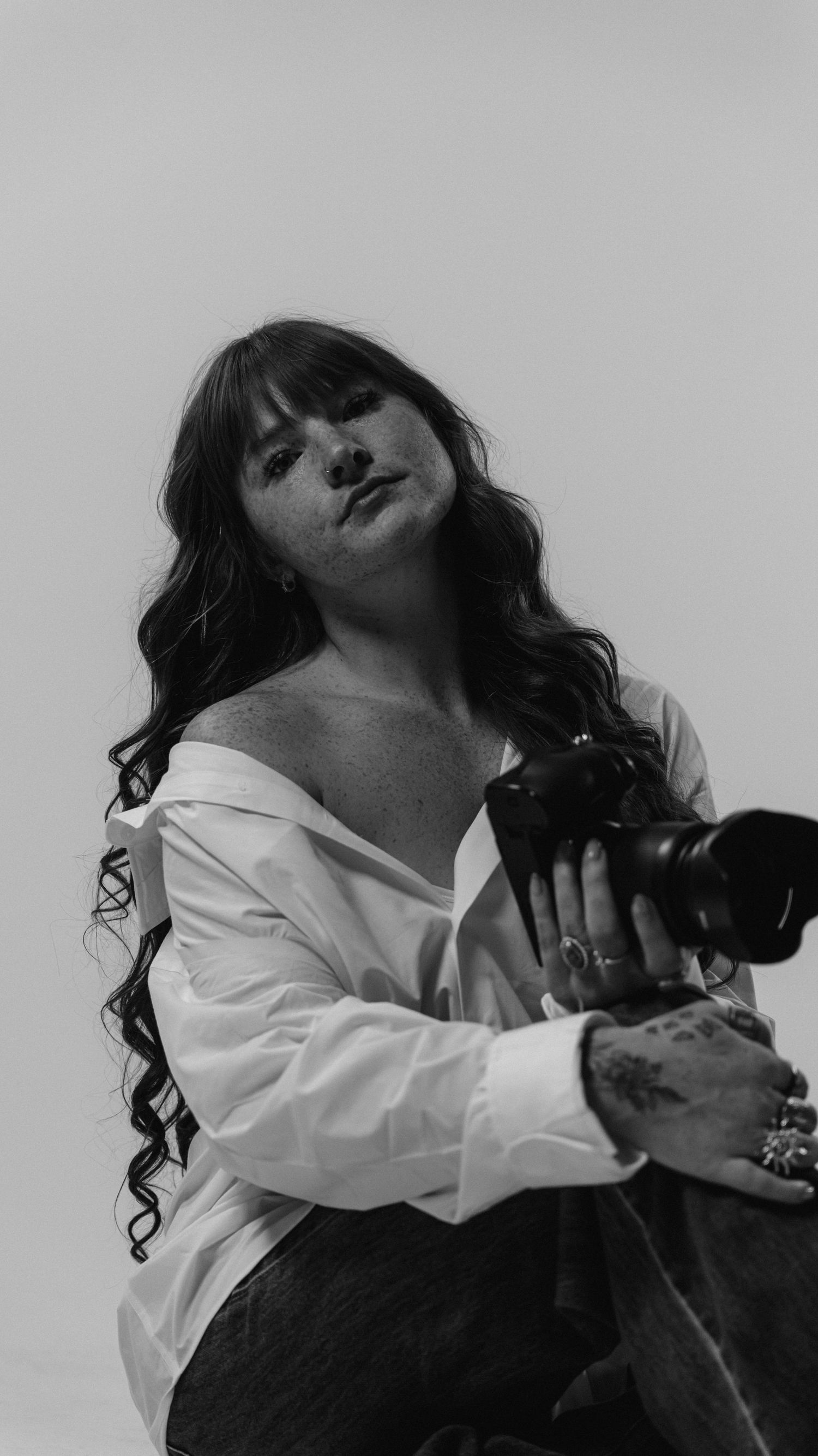 Black and white portrait of a woman with curly hair holding a camera, wearing an off-shoulder white shirt.