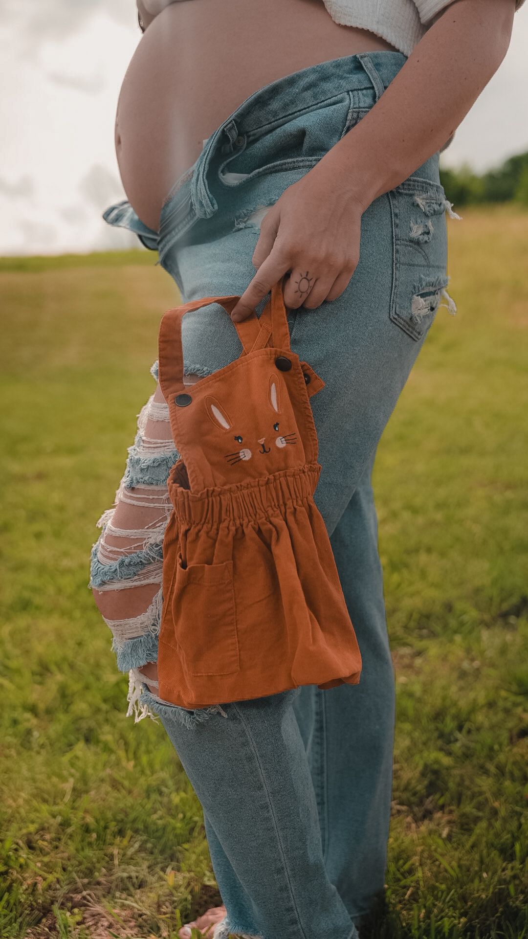Pregnant woman in ripped jeans holding a small orange bunny-shaped bag outdoors in a green field.