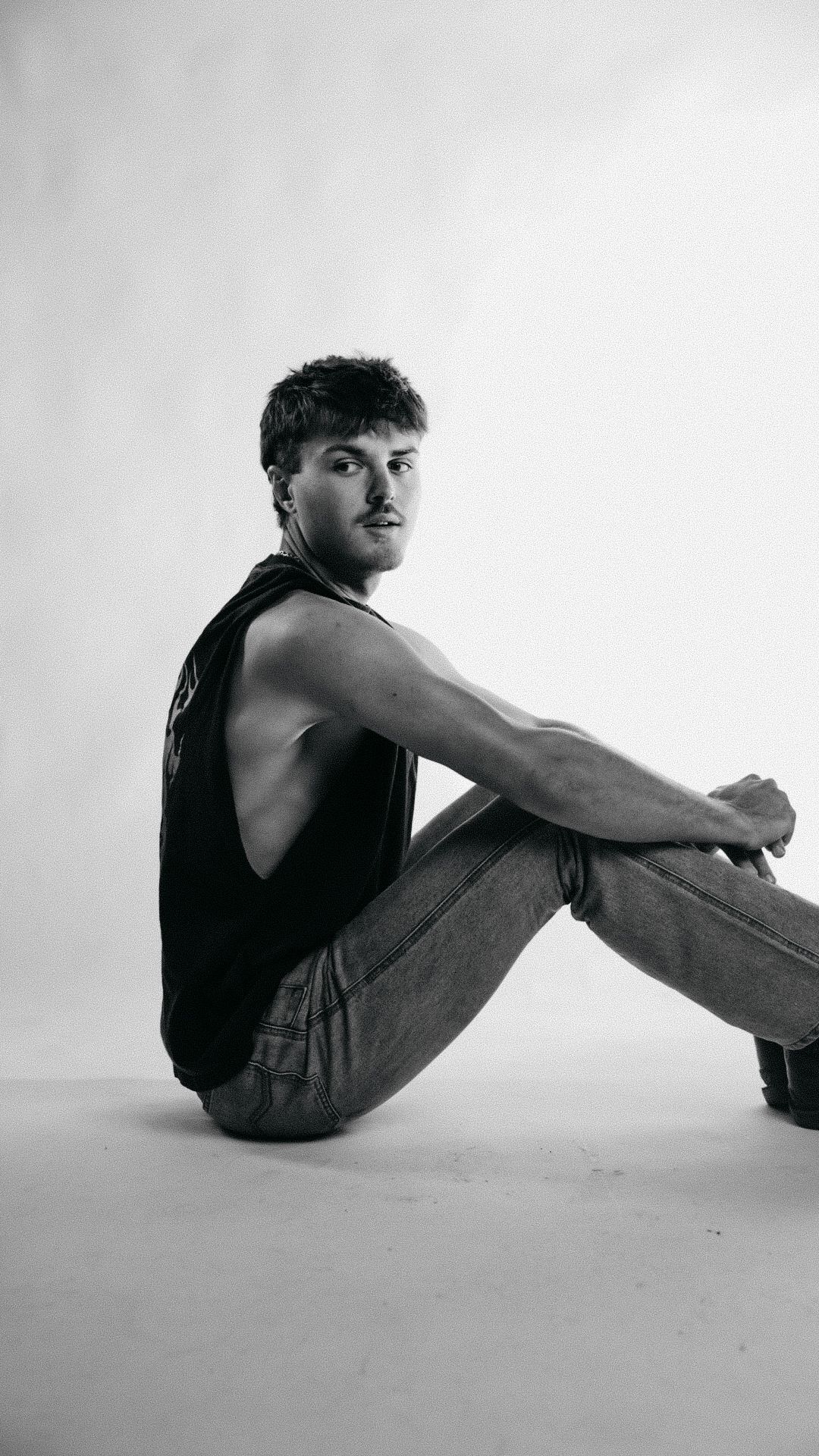 Black and white photo of a young man in a tank top and jeans sitting on the floor against a white background.