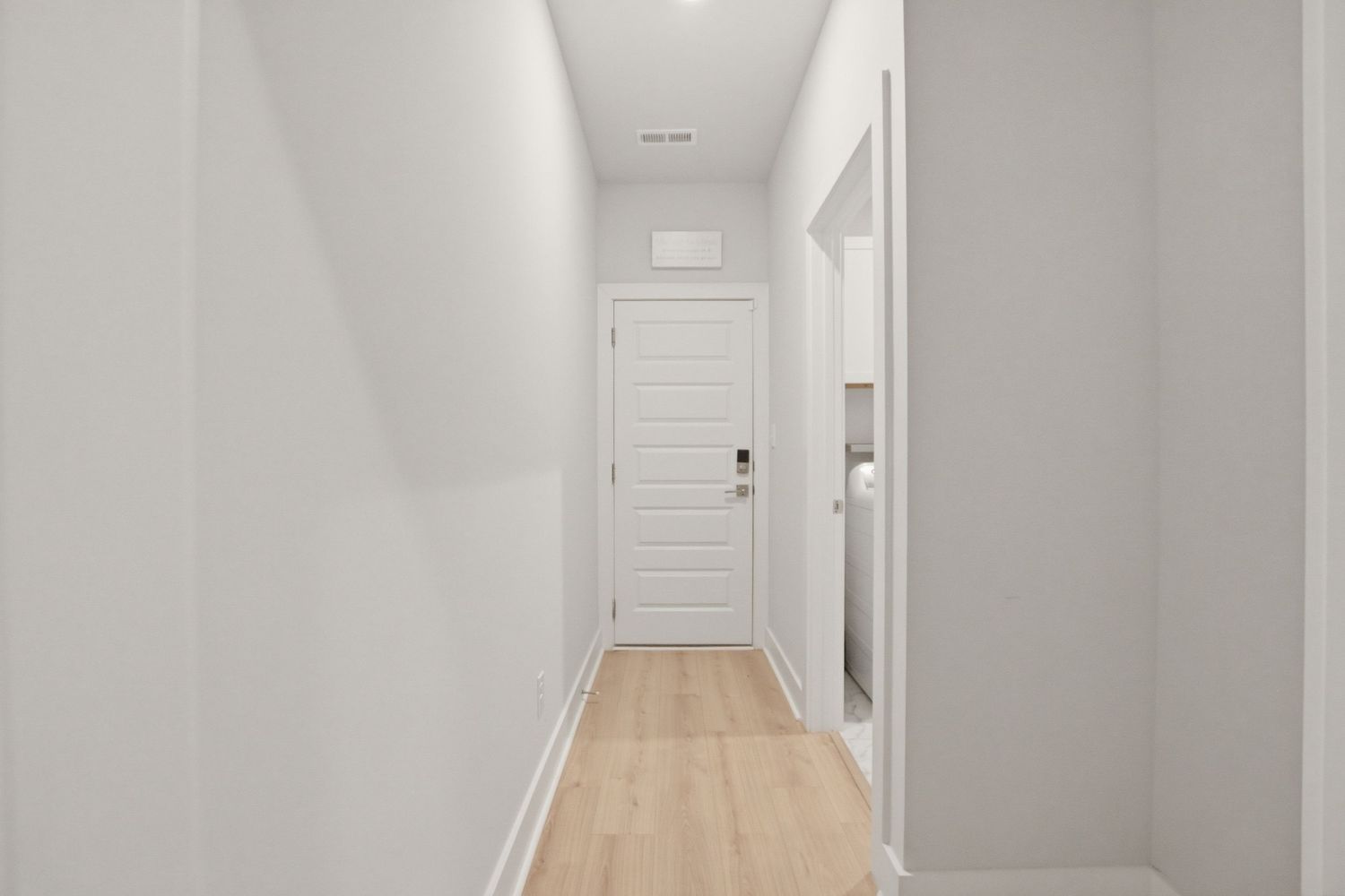 Narrow white hallway with light hardwood floors leading to a white paneled door.