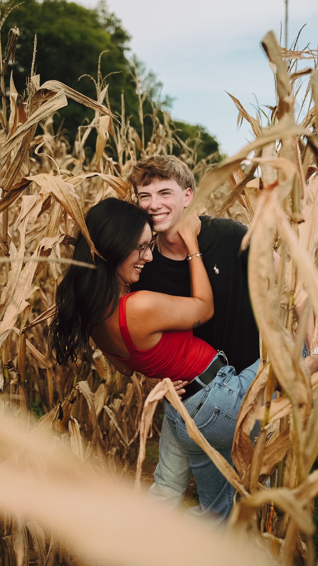 Happy couple embracing and laughing together in a golden autumn cornfield, woman in red top and man in black shirt.