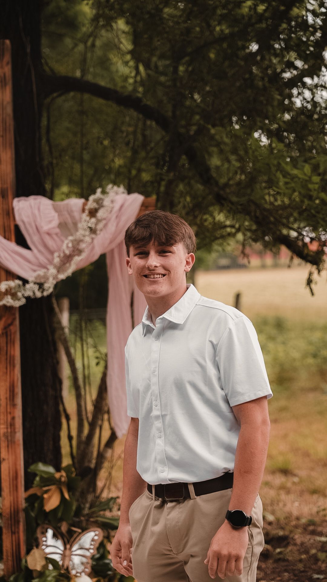 Smiling teenage boy in white button-up shirt and khaki pants standing outdoors near floral wedding arch.