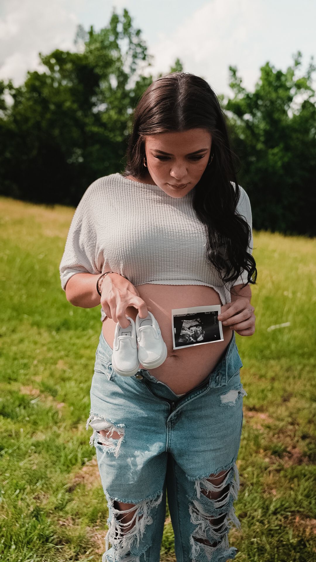 Expectant mother displaying ultrasound scan and tiny white shoes over her pregnant belly outdoors.