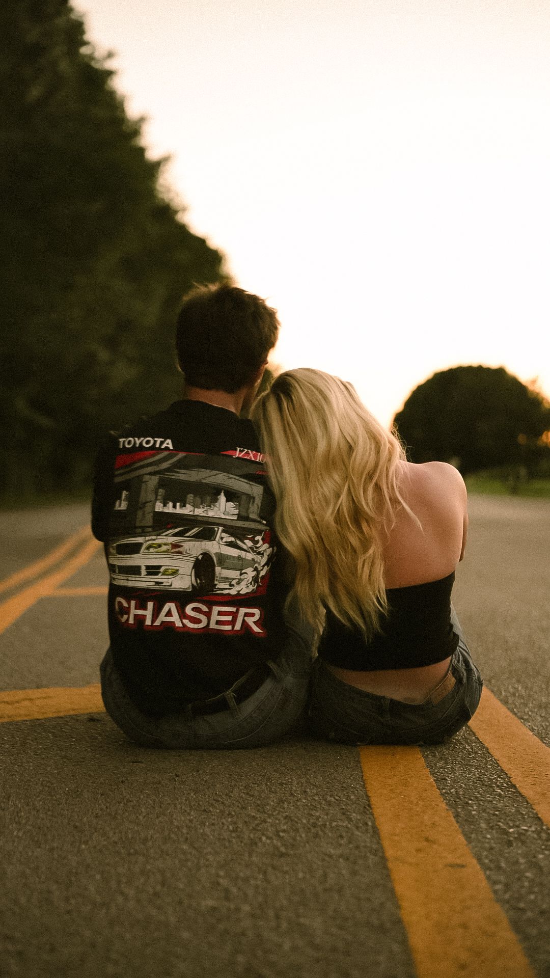 Couple sitting together on a road at sunset, man wearing a Toyota Chaser shirt, woman with blonde hair leaning on him.