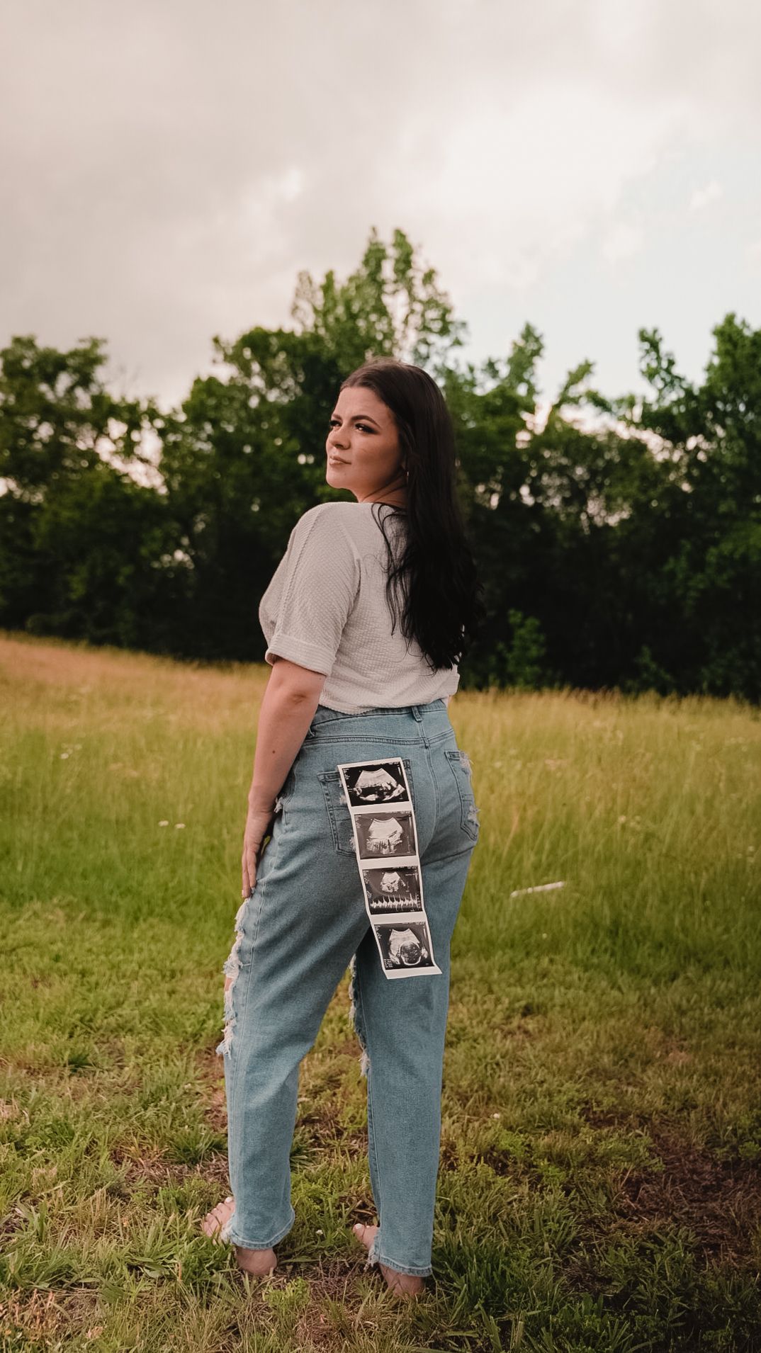 Pregnant woman in jeans displaying ultrasound photos on back pocket, standing in green field.