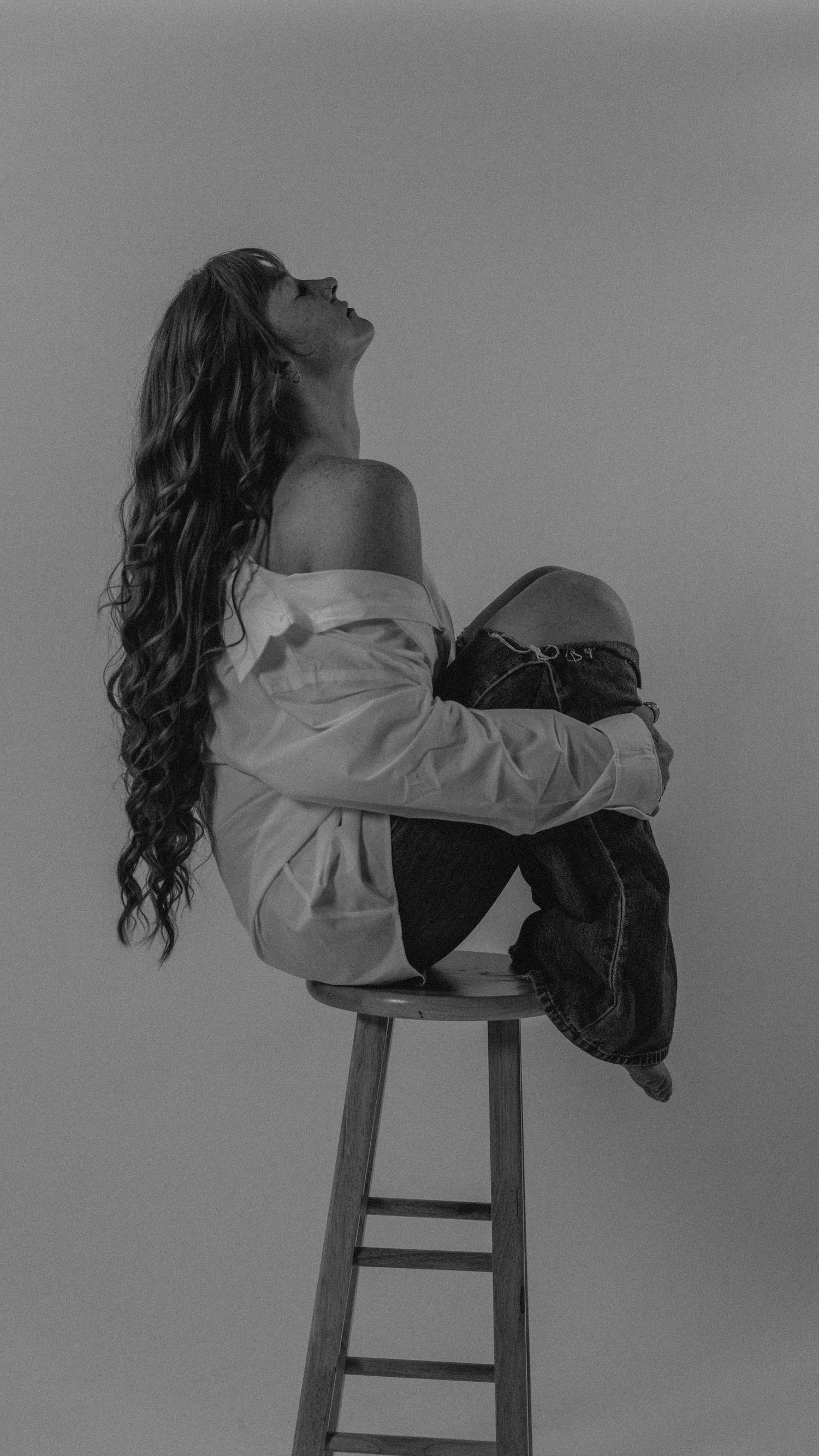 Black and white photo of woman perched on wooden stool in white off-shoulder shirt and jeans.