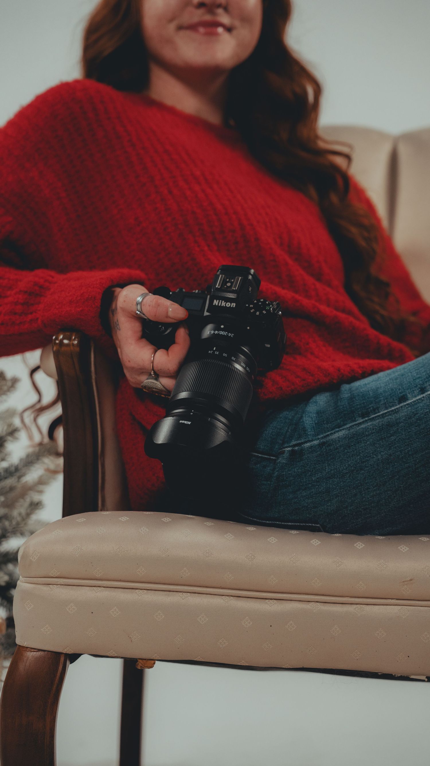 Woman in red sweater and jeans sitting in chair holding a Nikon camera, smiling.