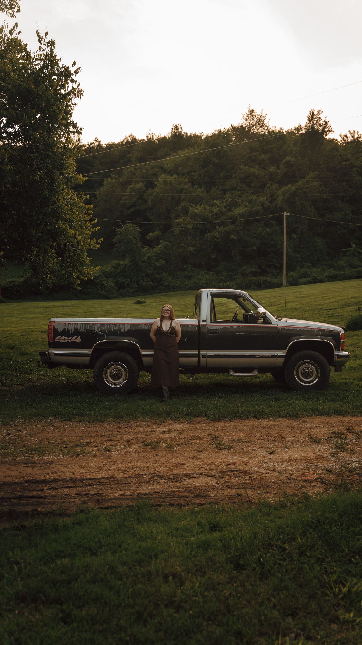 A woman stands beside a classic Chevy pickup truck on a rural dirt path surrounded by lush green hills at sunset.