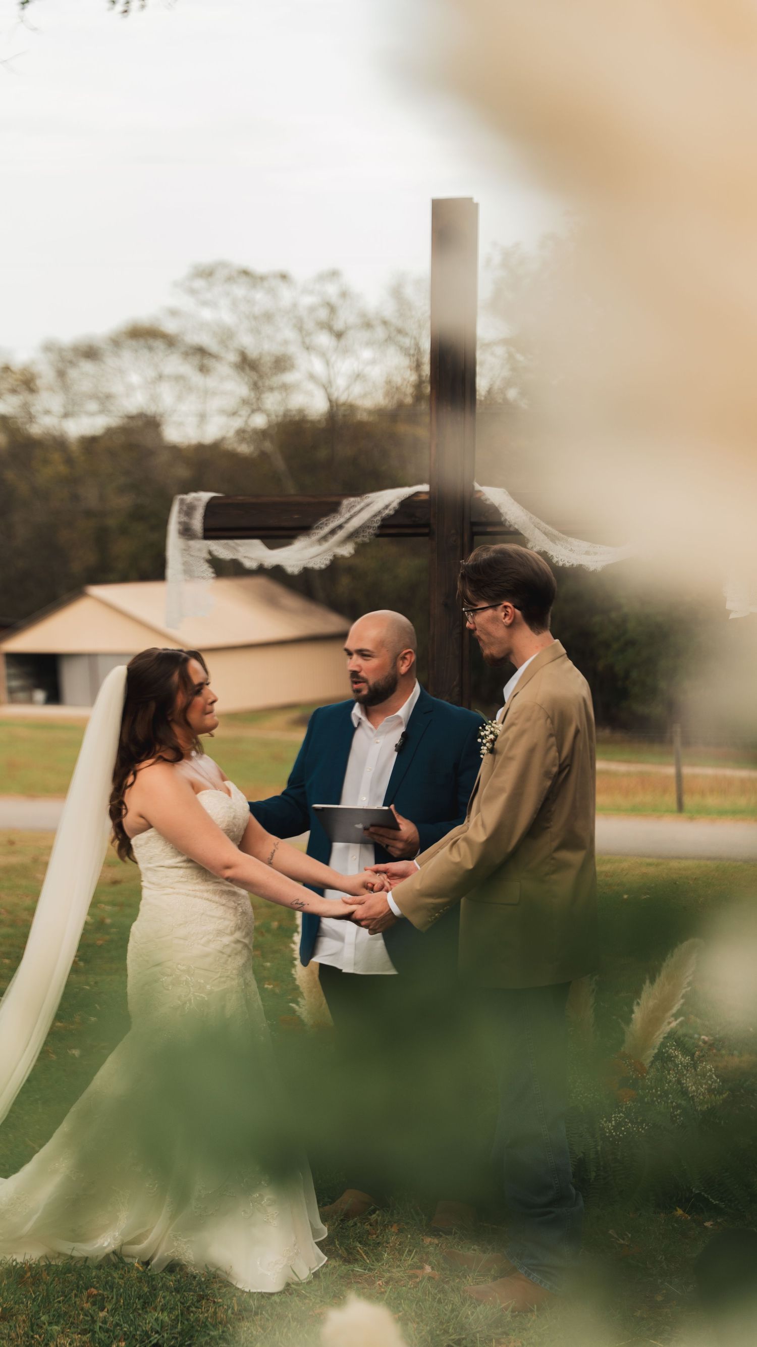 Outdoor wedding ceremony with couple exchanging vows before wooden cross with officiant present.