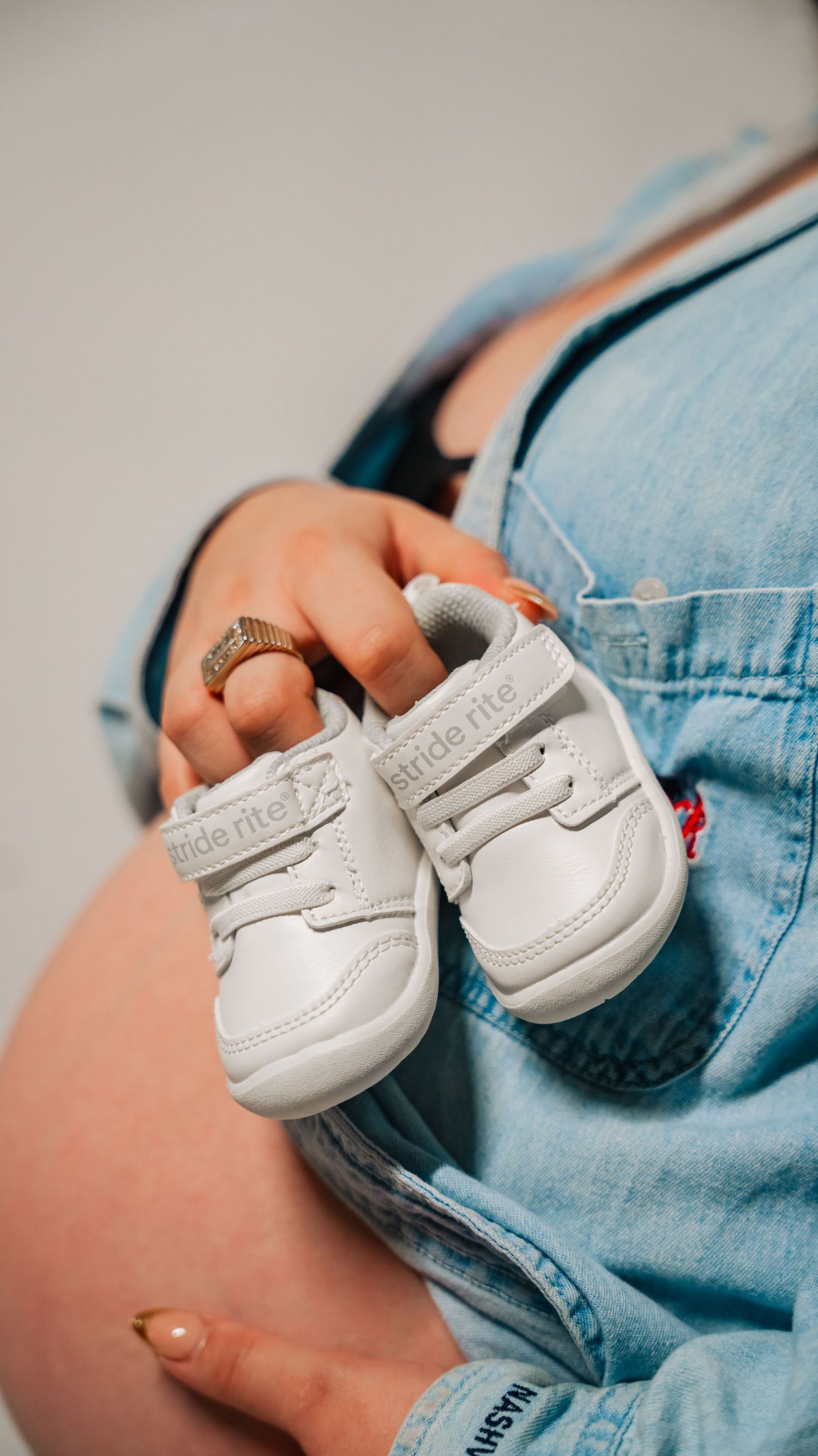 Pregnant woman in denim overalls holding tiny white Stride Rite baby sneakers against her baby bump.