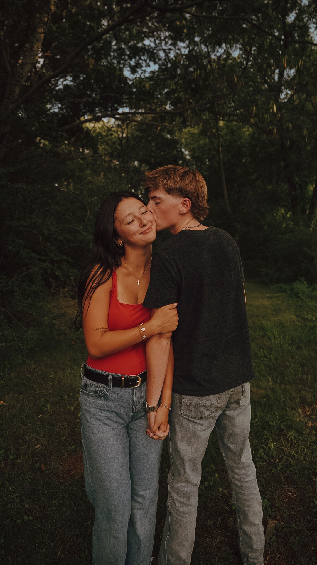 A young couple shares a tender kiss in a lush green park, the woman in a red top and jeans smiling warmly.