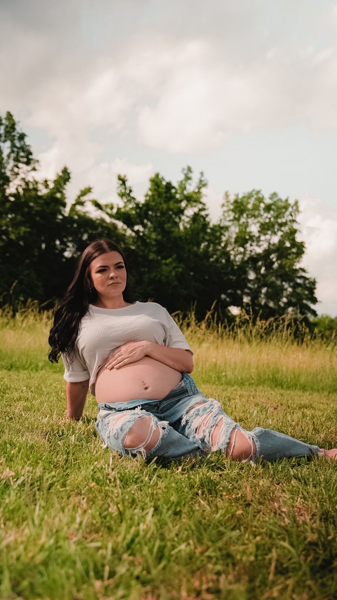 Pregnant woman in ripped jeans and white top sitting on grass in outdoor maternity photo session.