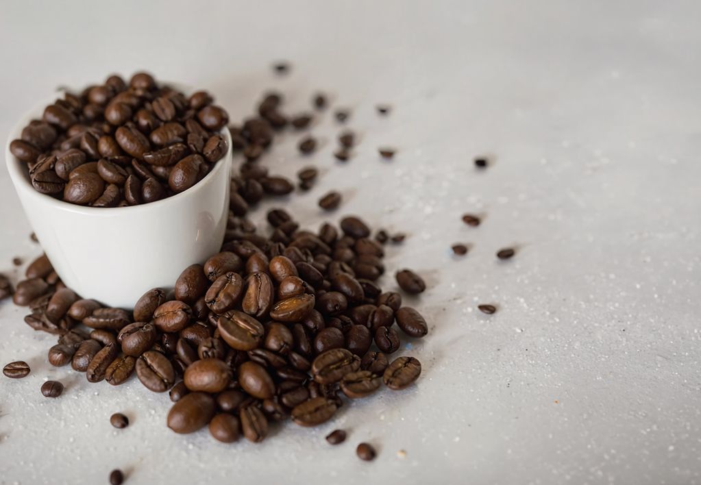 Minimalist close-up of a white cup overflowing with roasted coffee beans, with more beans scattered across a light stone surface.