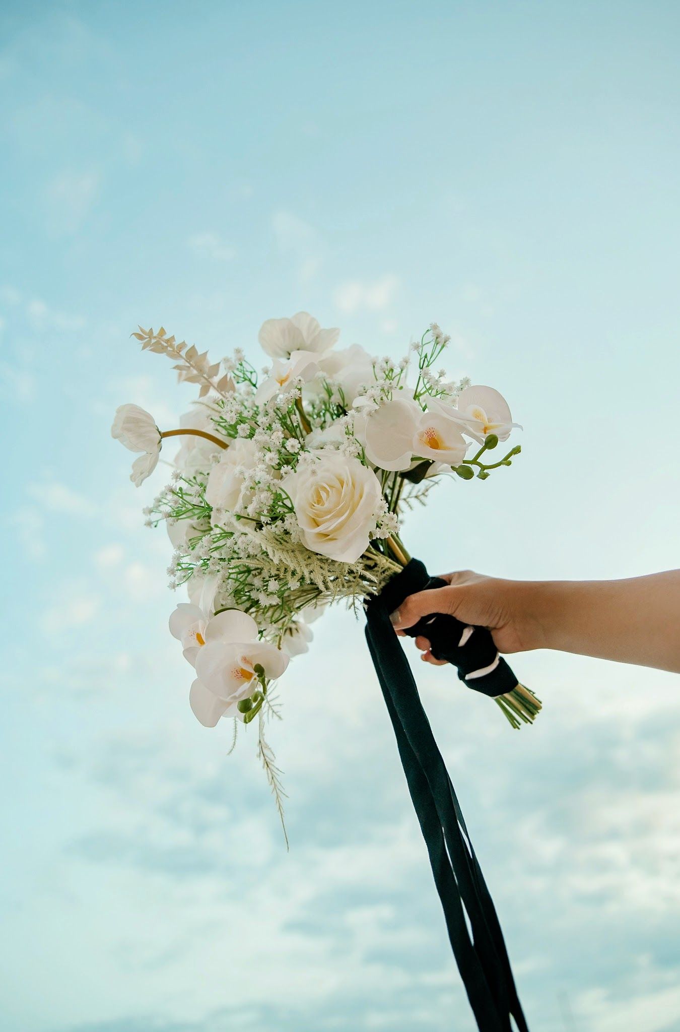 White rose and orchid bouquet held against light blue sky.