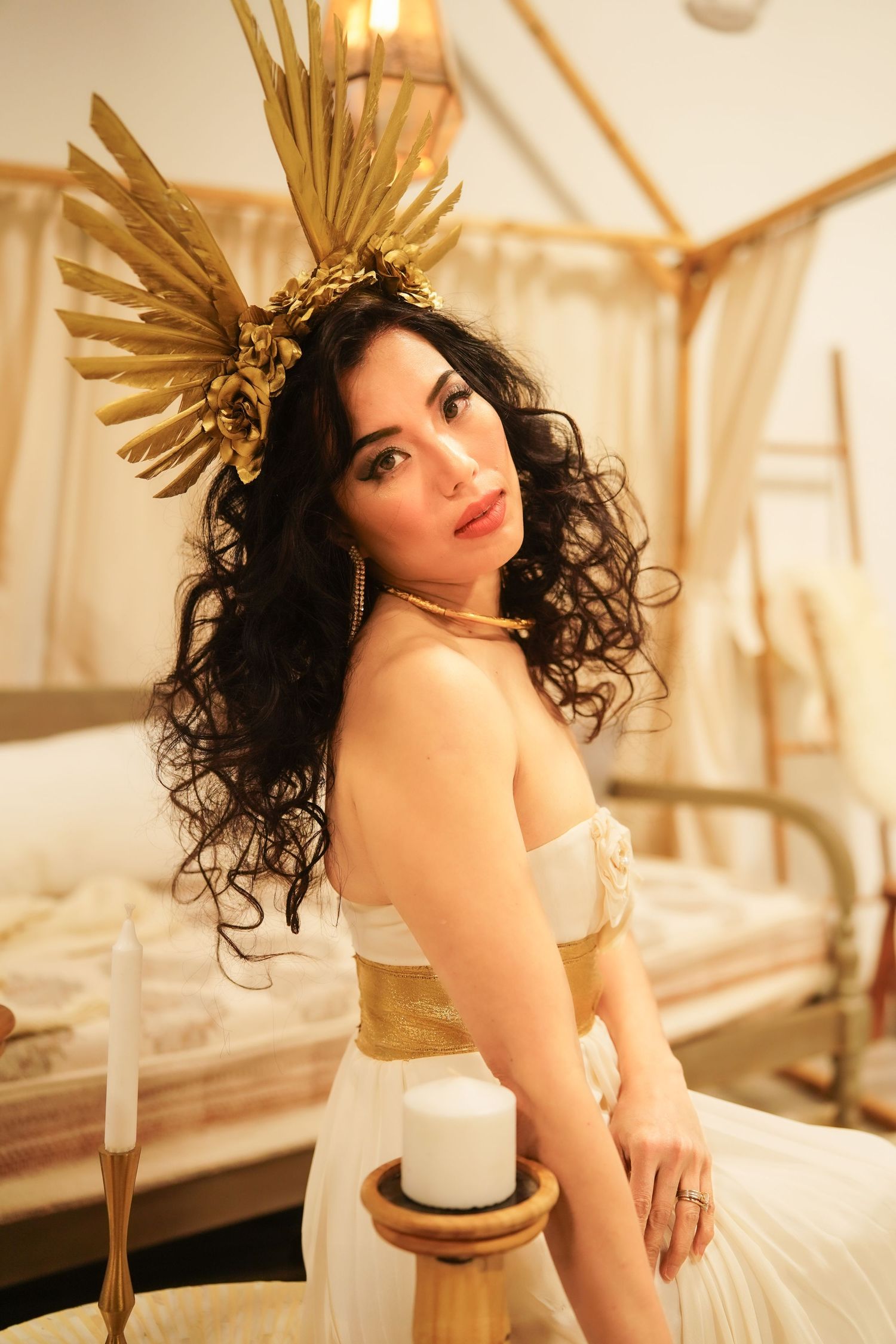 A figure wearing a golden crown headpiece and white dress poses in a dreamy indoor setting with warm lighting.