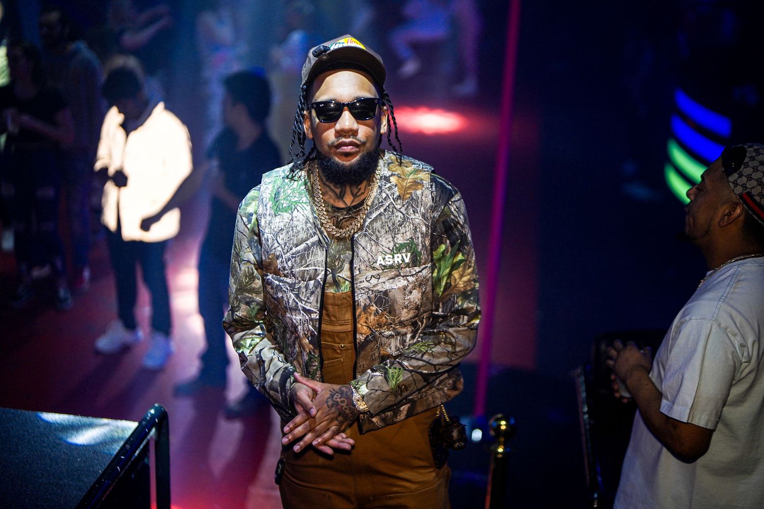 A person in sunglasses and shiny metallic jacket poses at a nightclub with blue lighting and dark atmosphere.