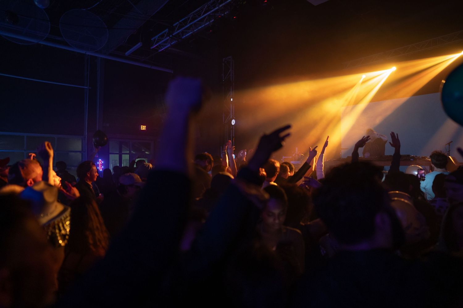 Concert crowd with raised arms illuminated by warm golden spotlights in dark venue.