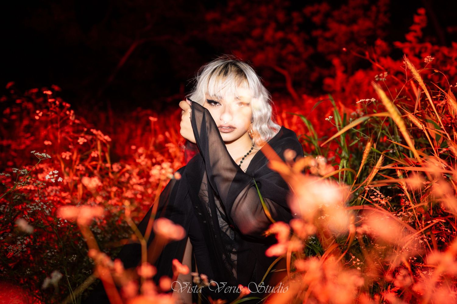 Fashion portrait amid red-tinted foliage with dramatic lighting and sheer black attire.