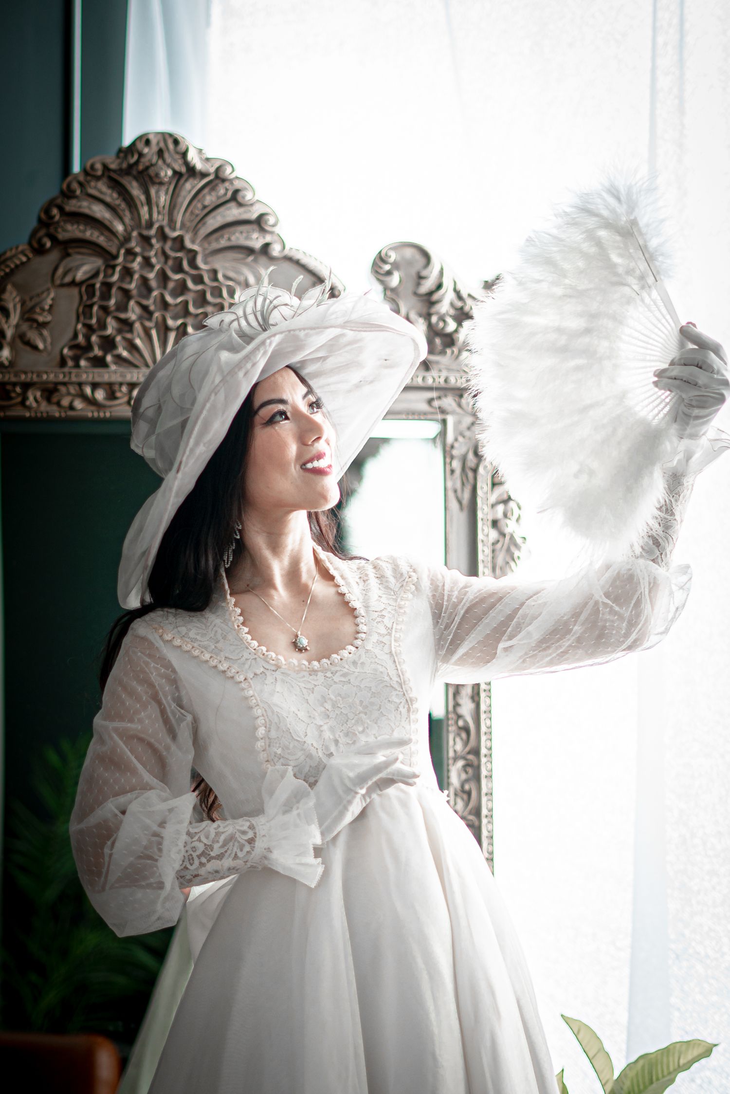 Bride in vintage white dress holding feather fan near ornate mirror.