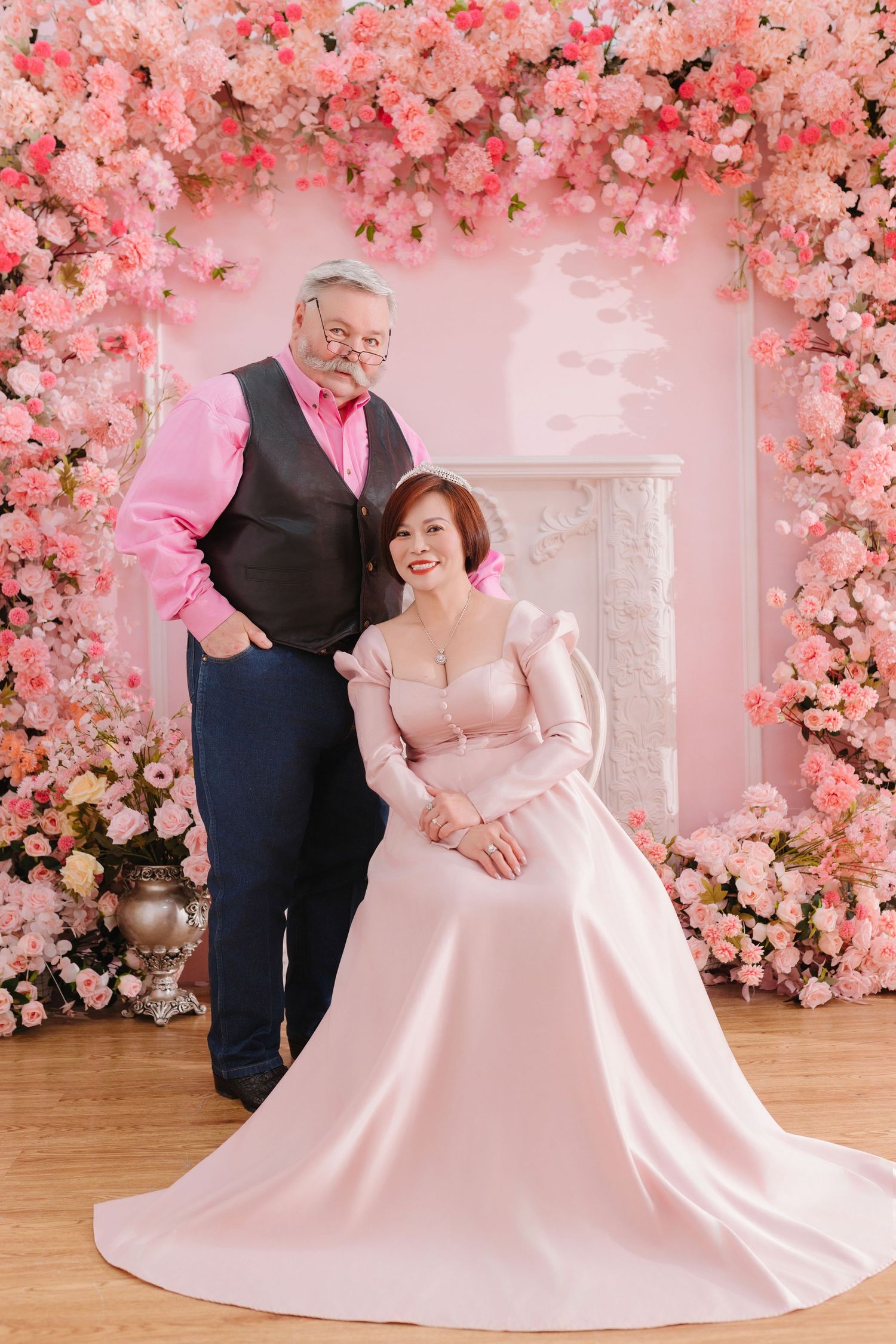 Wedding couple poses in front of romantic pink floral arch with cherry blossoms and elegant fireplace backdrop.