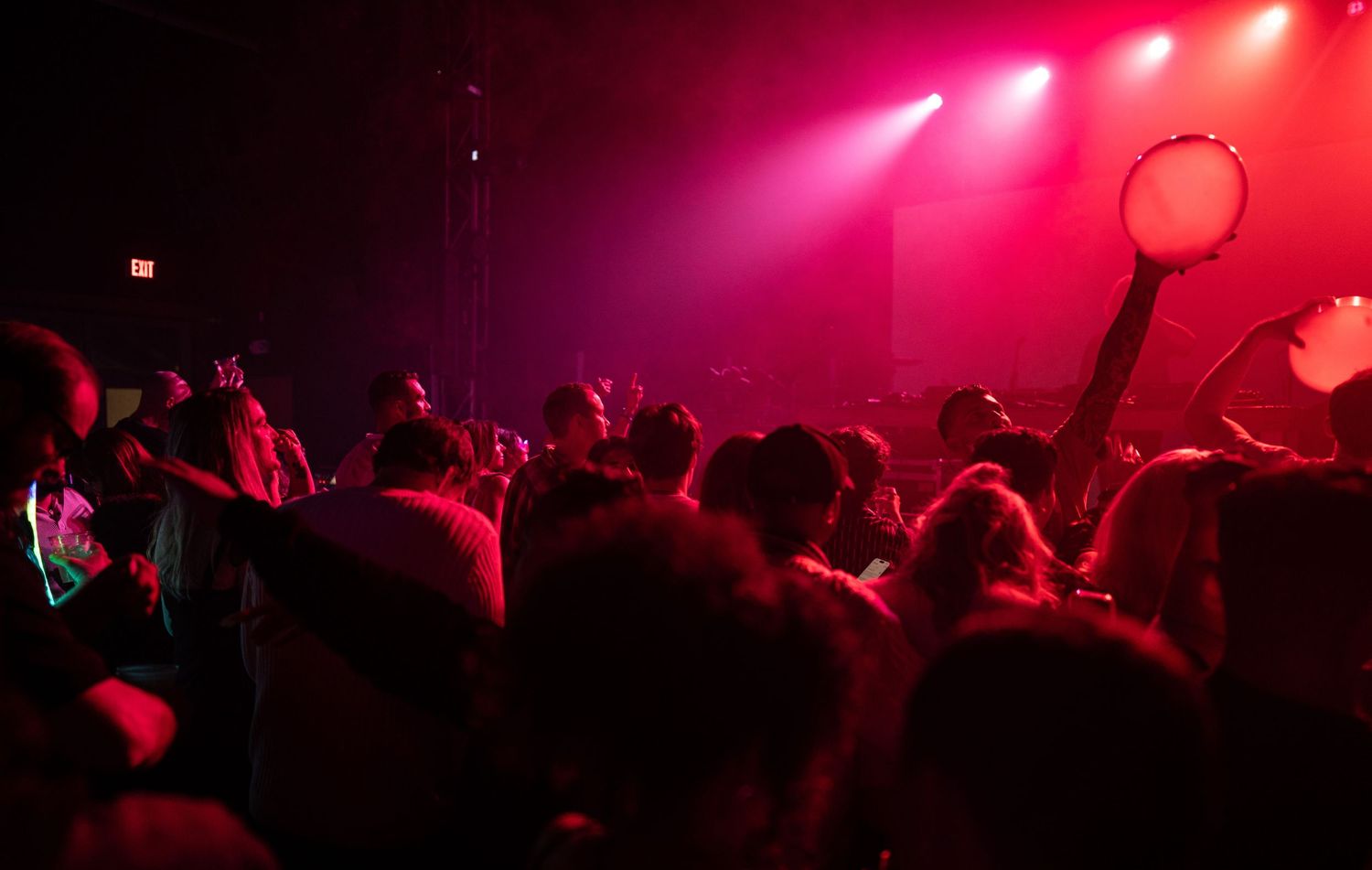 Concert crowd silhouettes gather under dramatic pink and red stage lighting during a live performance.
