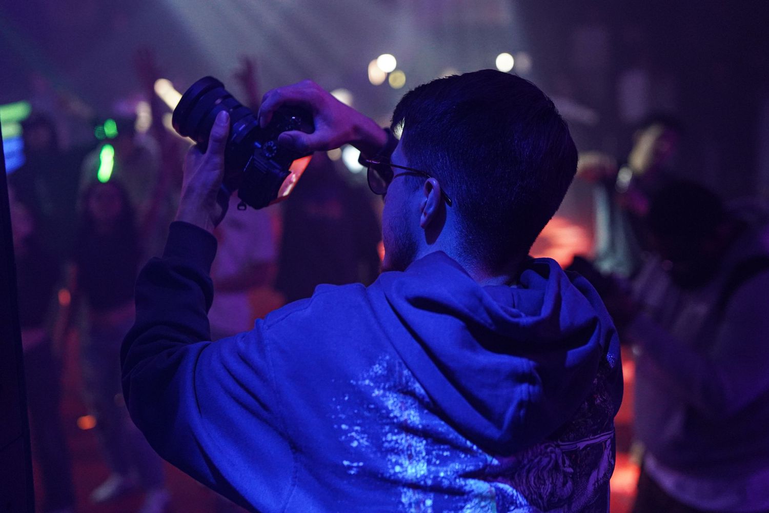 Someone in a blue hoodie holds up their phone at a dark concert venue with colorful lights visible in the background.