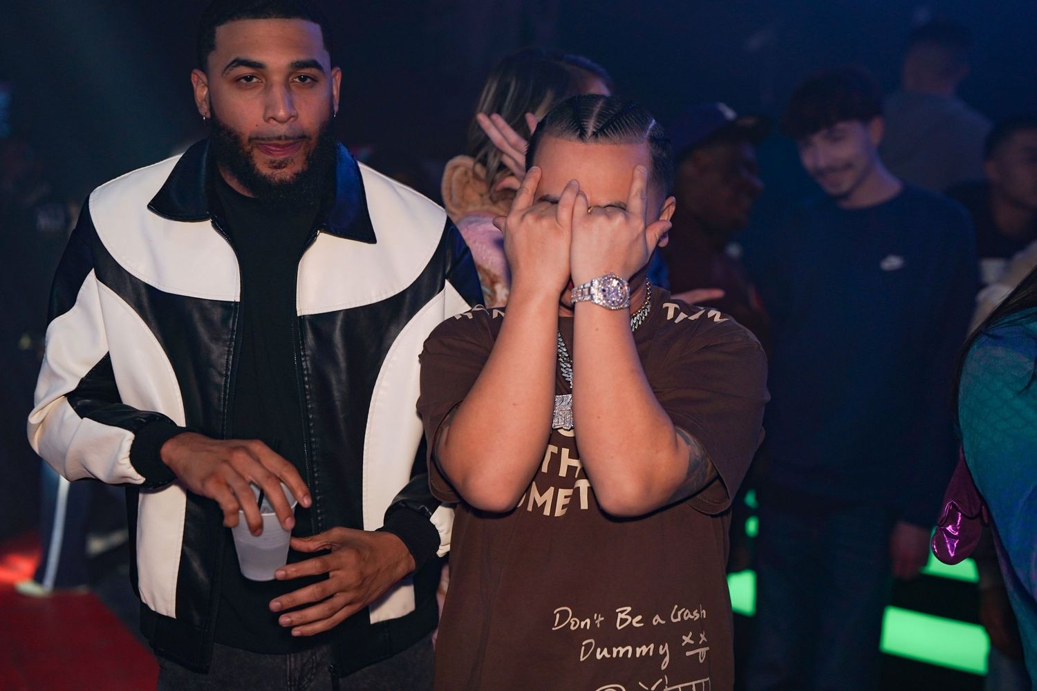 Two people at a nightclub venue with one wearing a black and white jacket and another in a brown t-shirt.