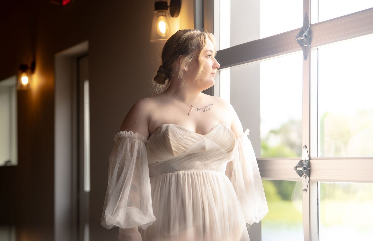 Elegant bride in off-shoulder gown gazes out window in soft light. At Wichita, Kansas.