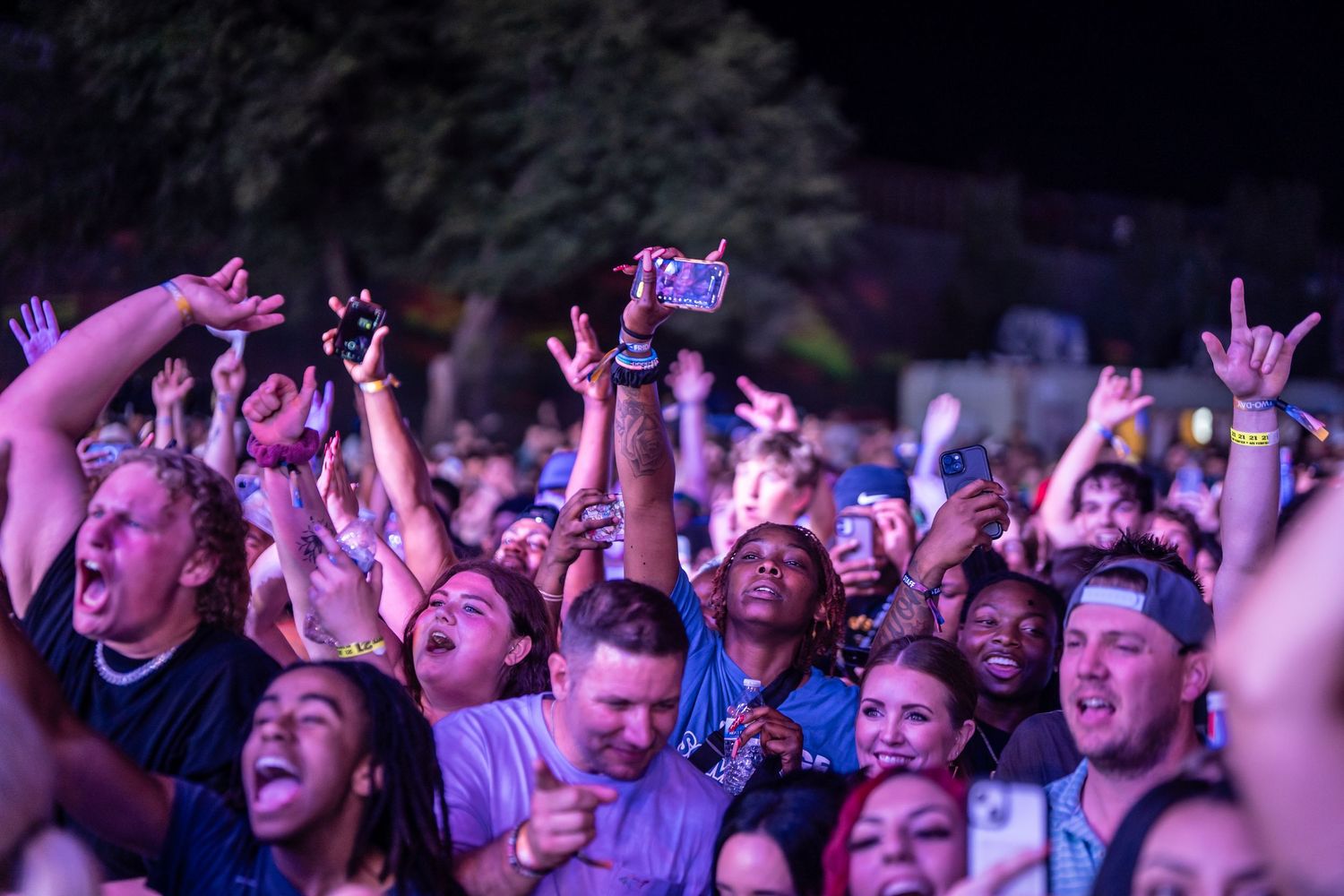 Enthusiastic concert crowd raises hands and cheers during a live music performance under purple lighting.