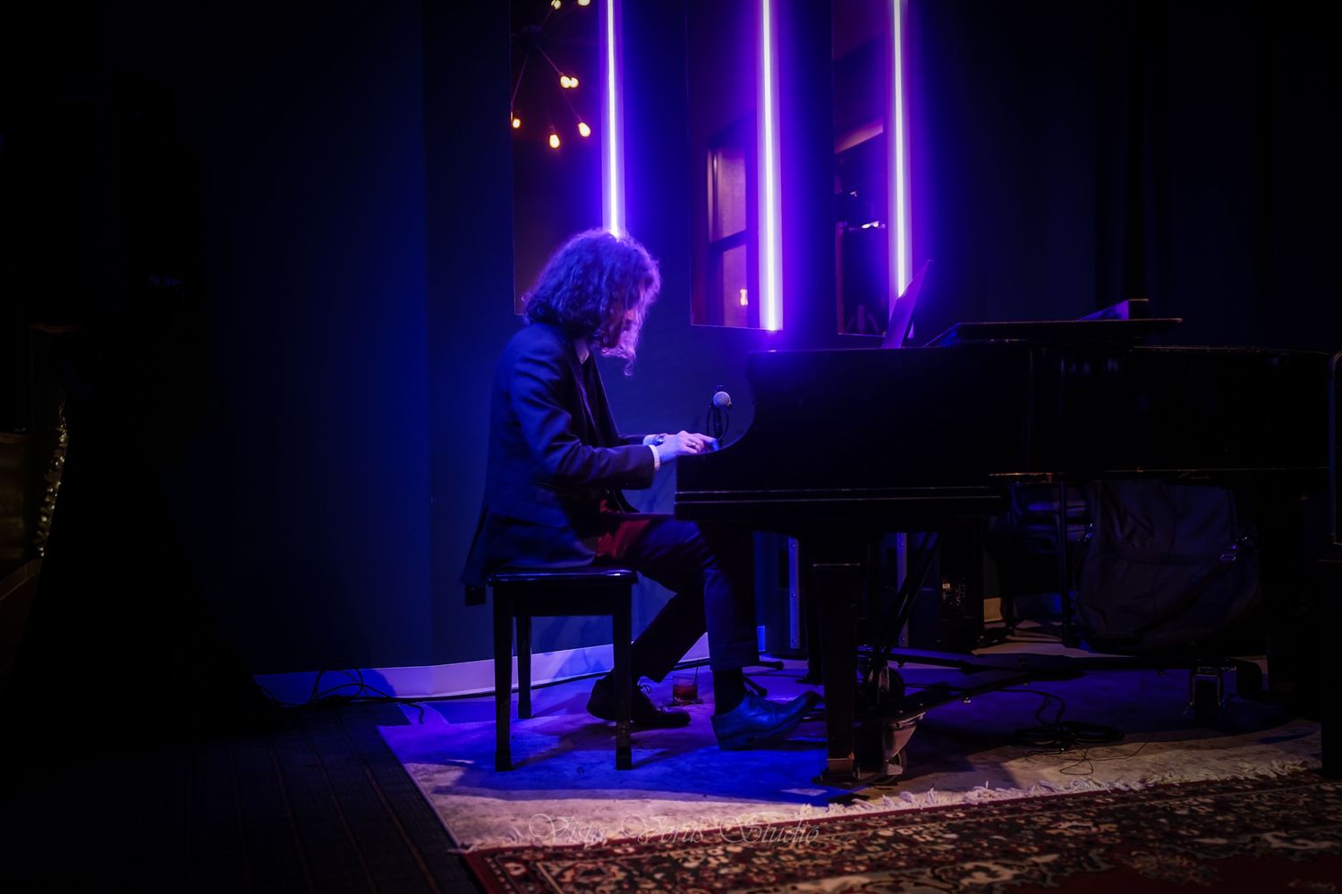 Pianist performing on grand piano under dramatic purple stage lighting in dark venue.
