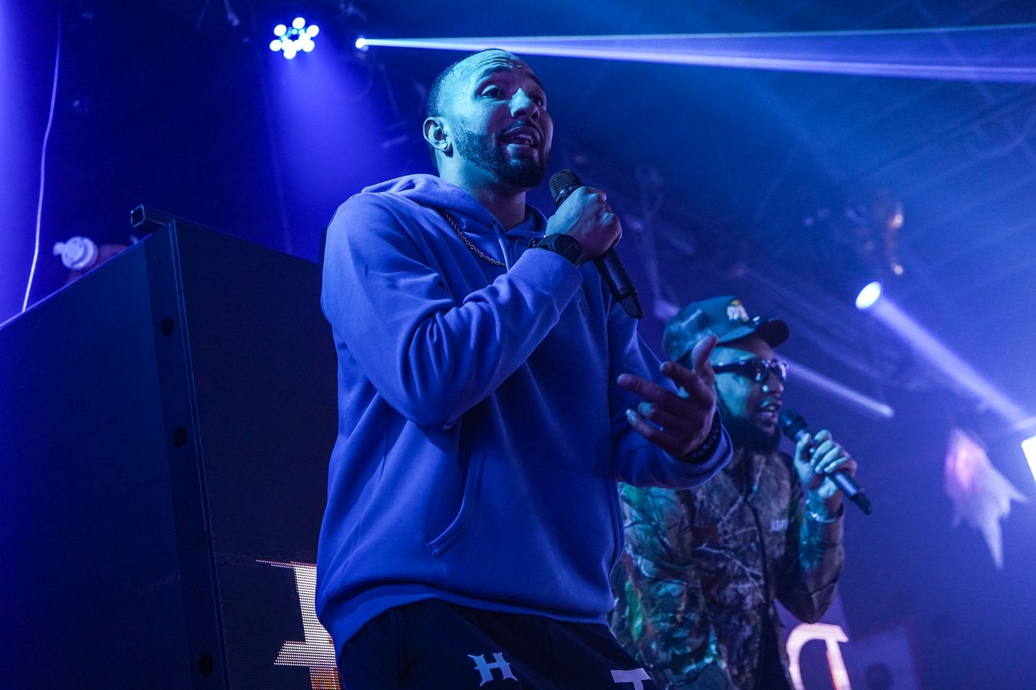 A performer in a blue sweatshirt sings passionately on stage under dramatic blue lighting at a concert performance.