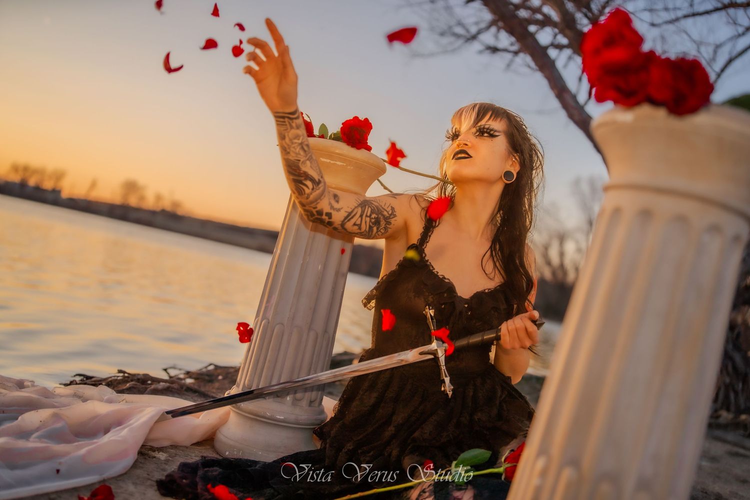 Person in vintage dress tossing red rose petals by water during sunset.