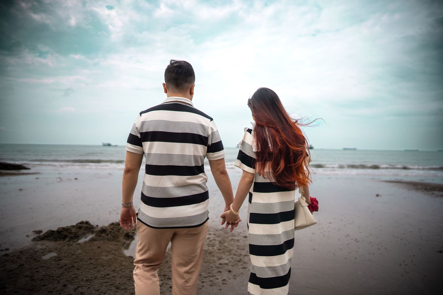 Two people walk together on a sandy beach near the ocean under a cloudy sky at sunset.