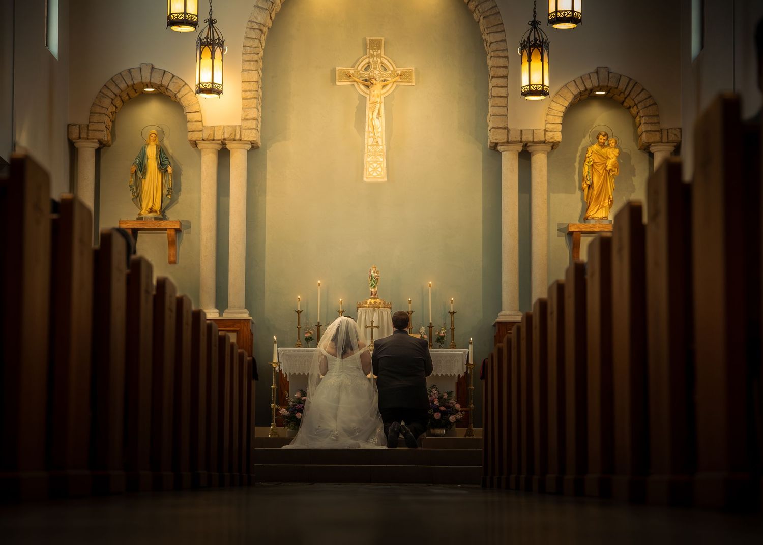 Dimly lit Catholic church interior with wooden pews and illuminated statues and crucifix in golden lighting.