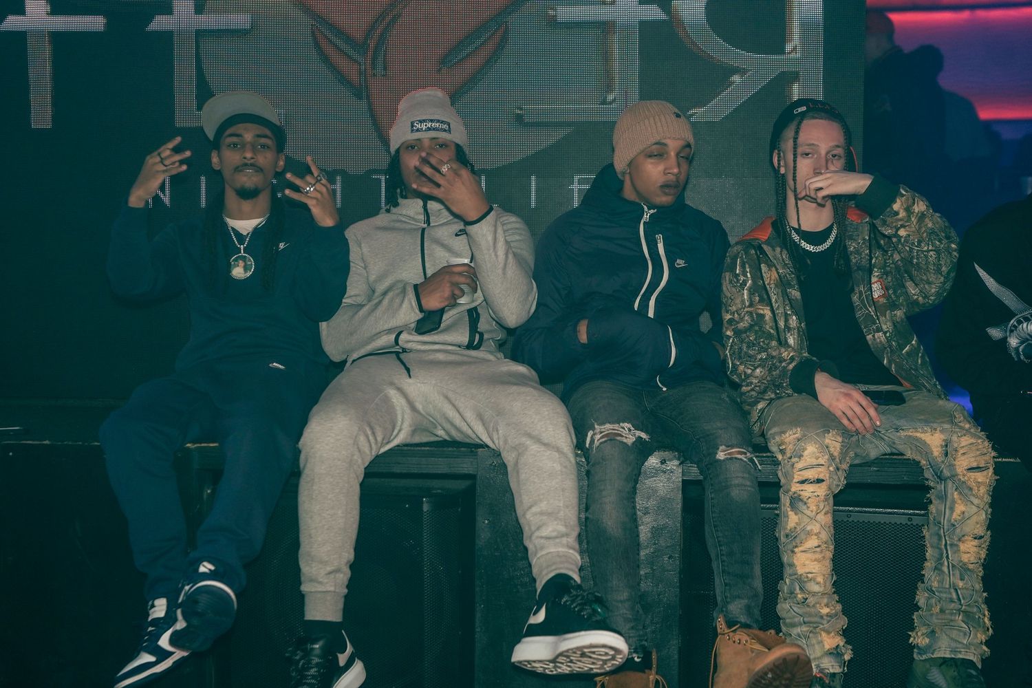 Four people sit together in casual streetwear clothing on a dark stage with colorful lighting in the background.