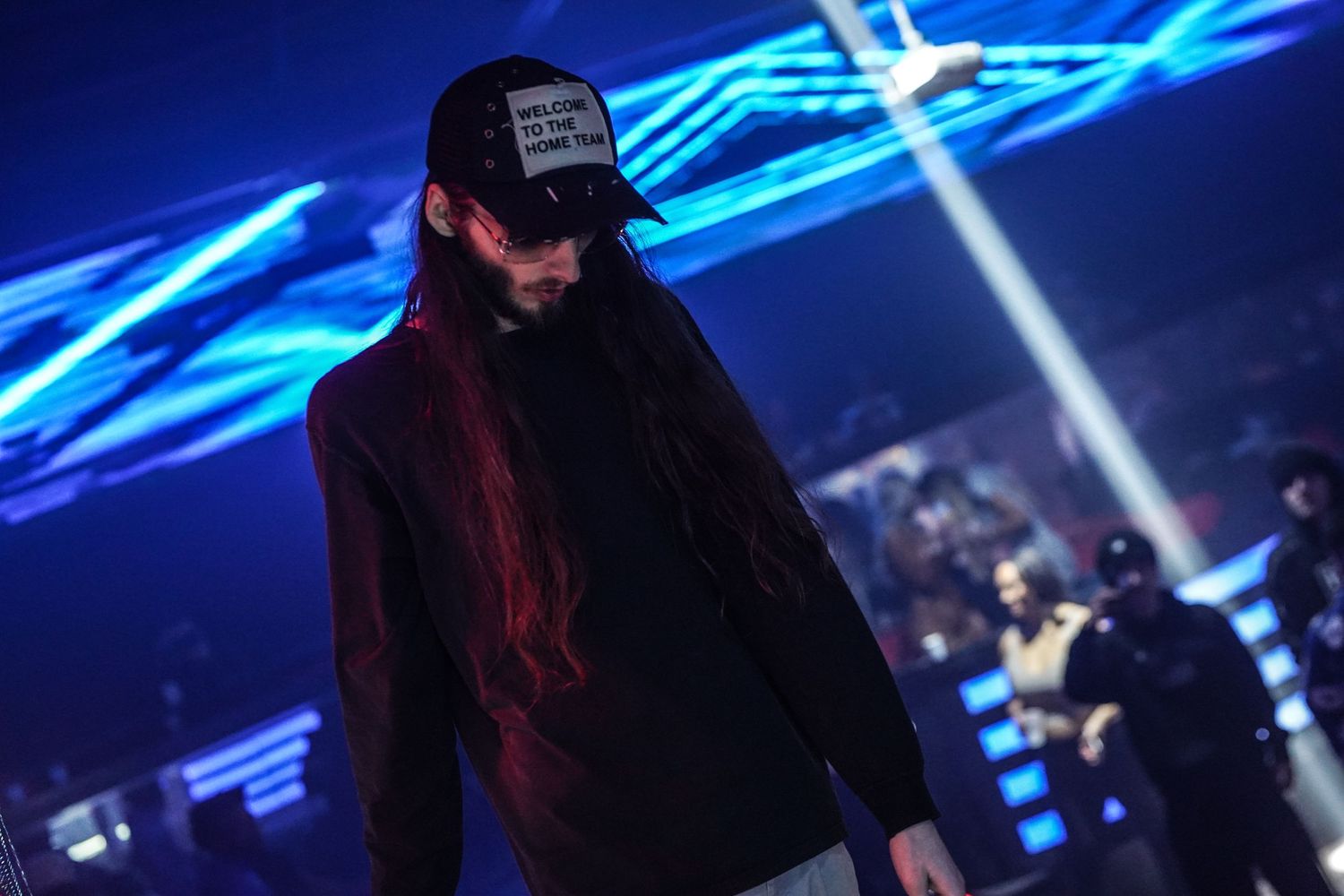 A person in black clothing performs on stage with blue neon lights and laser beams creating a dramatic atmosphere.
