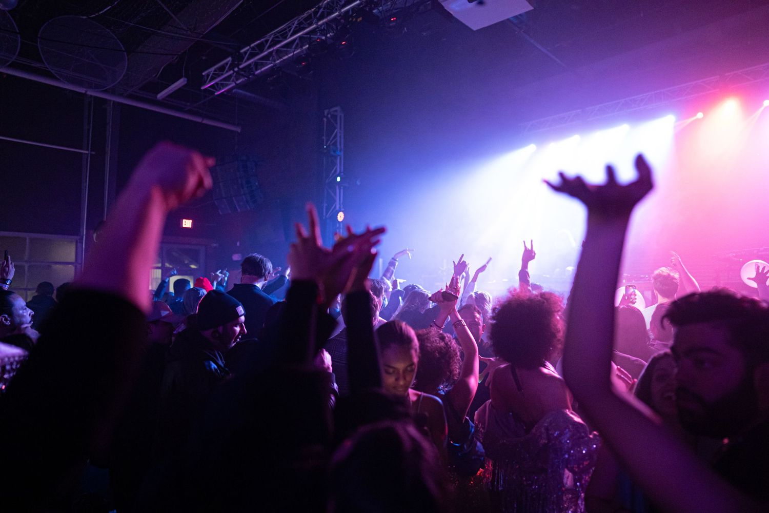 Silhouetted crowd raises hands at a concert with purple and pink stage lighting.