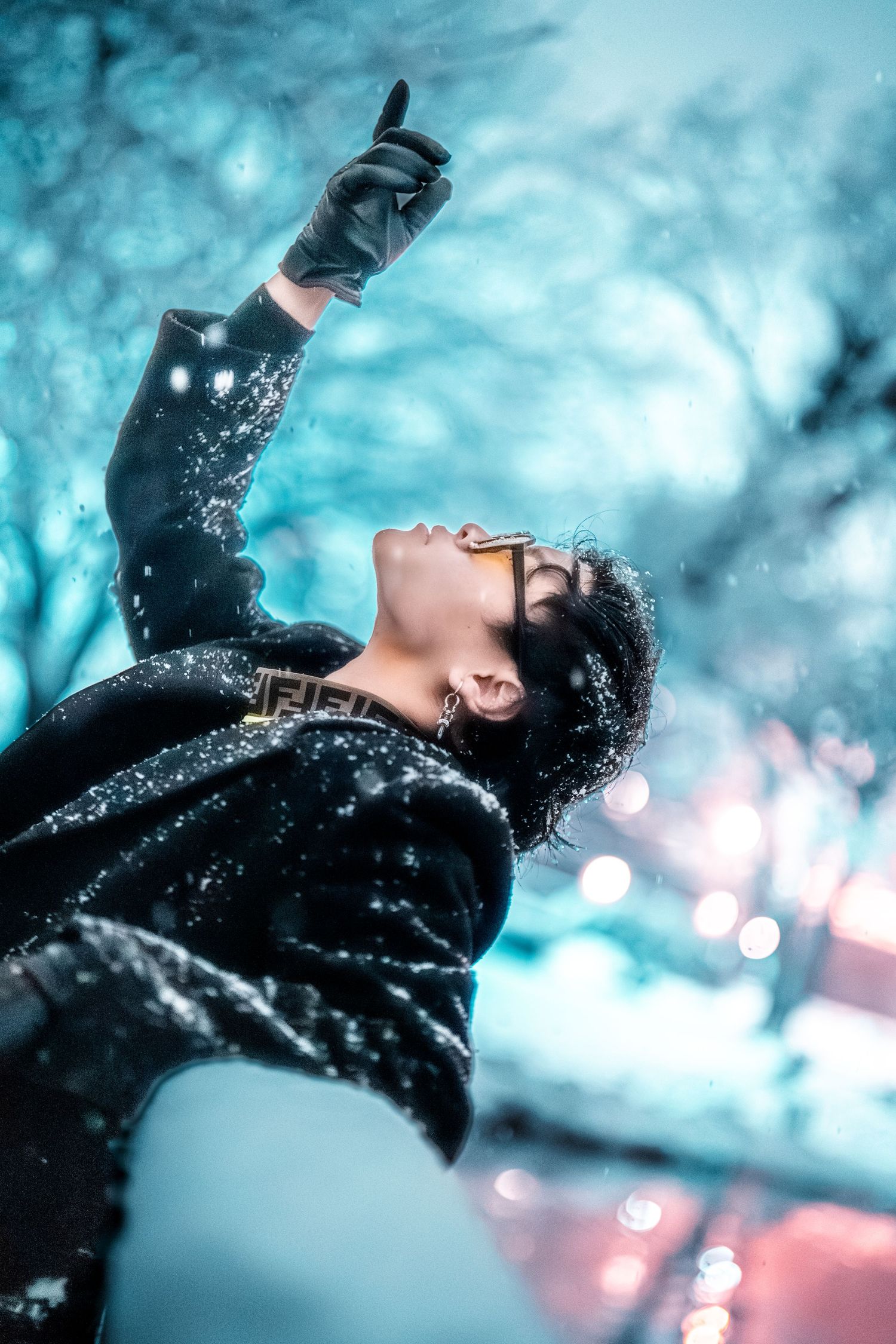 Person wearing black gloves reaches upward against snowy turquoise winter backdrop with dreamy bokeh lights.