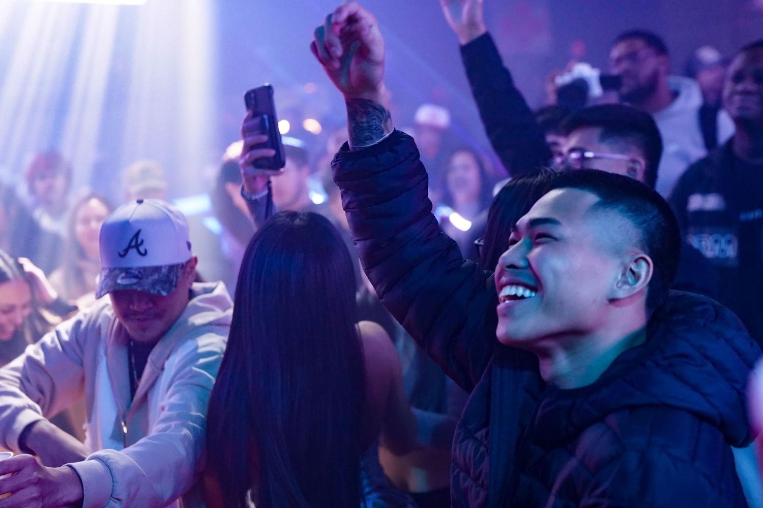 Crowd of enthusiastic people dancing and cheering at a nightclub with purple lighting effects in the background.