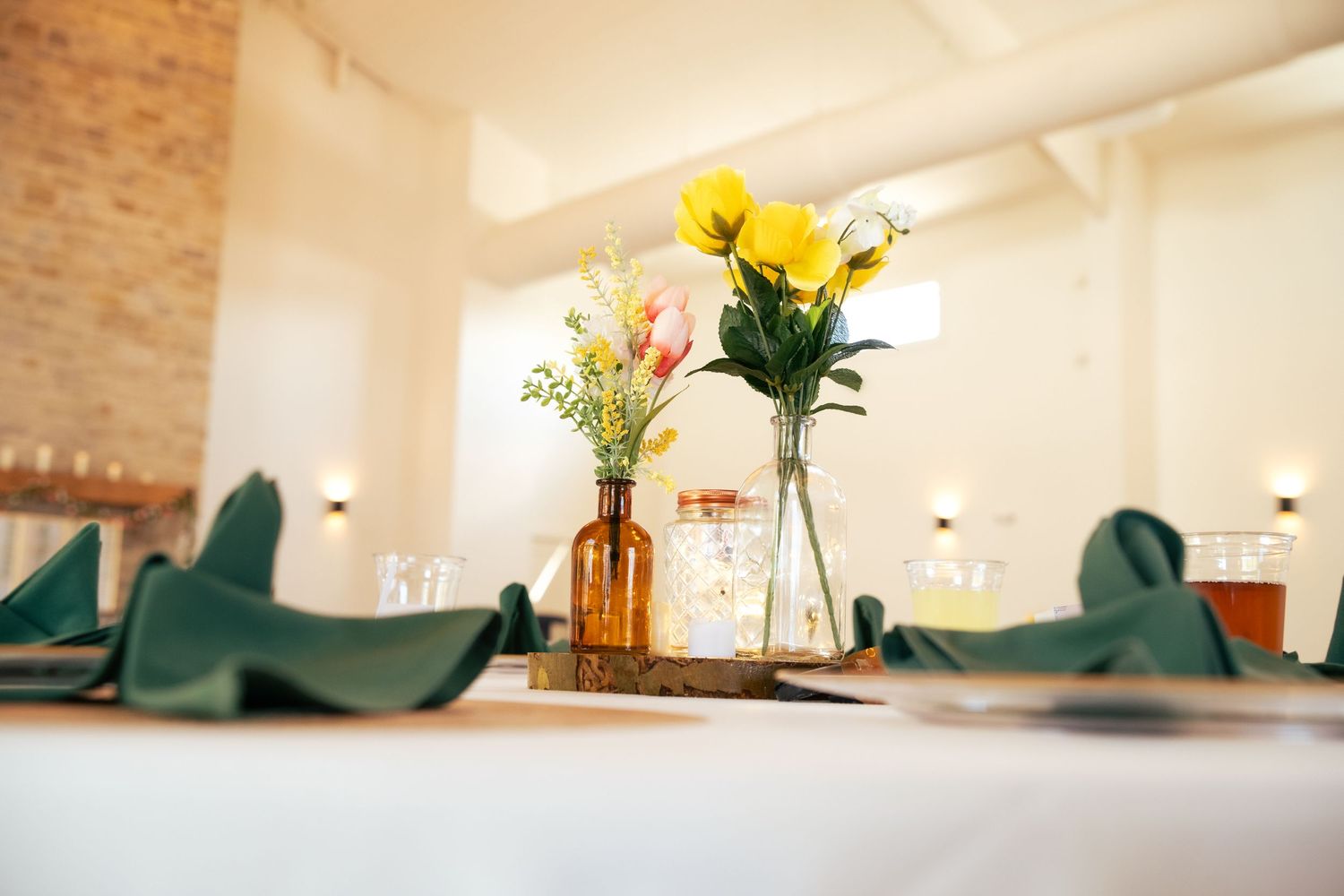 Yellow and pink flowers in amber vases with folded napkins on a wedding reception table.
