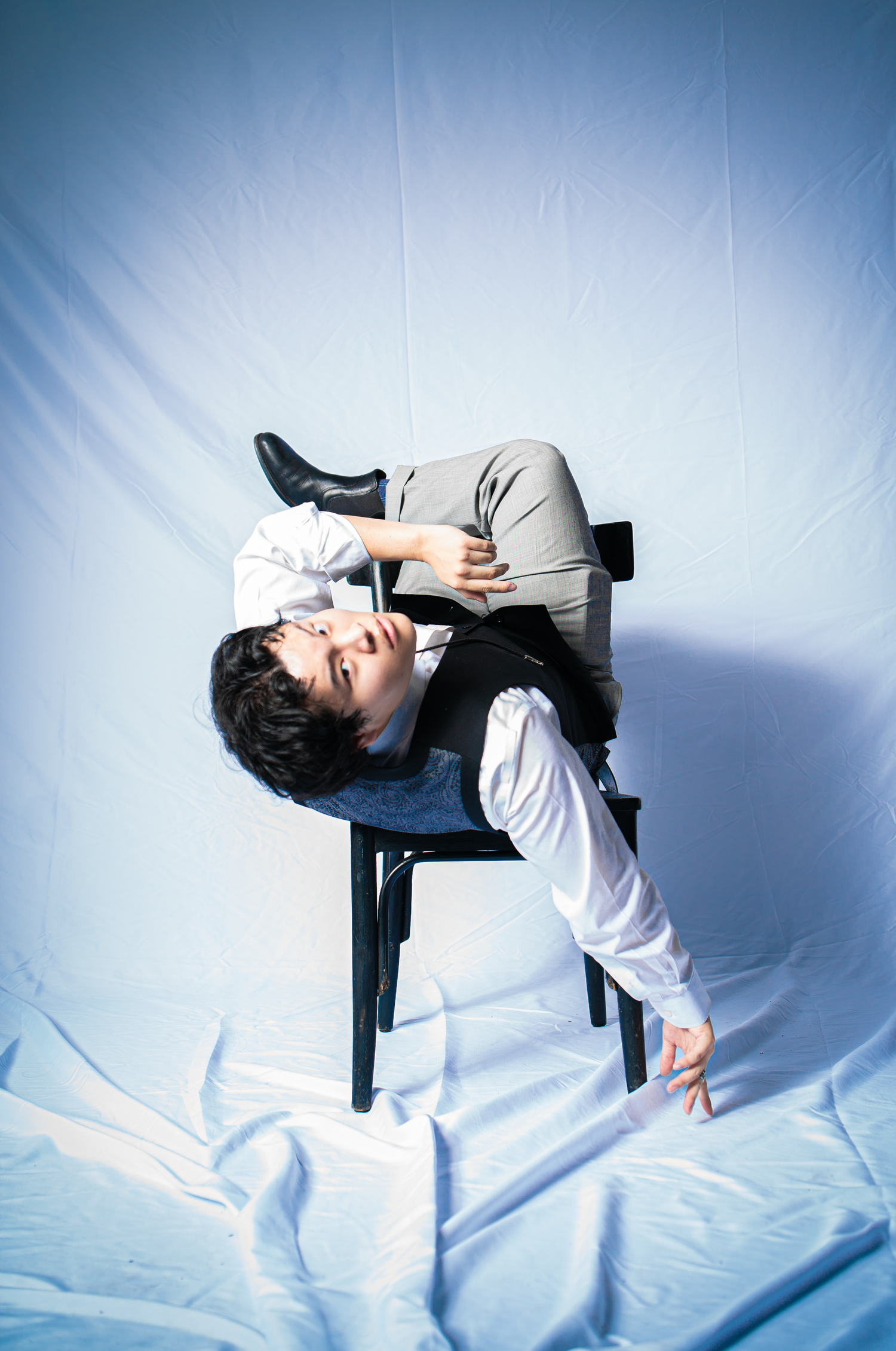 Person in casual attire lying sideways on a black chair against blue backdrop in artistic pose.