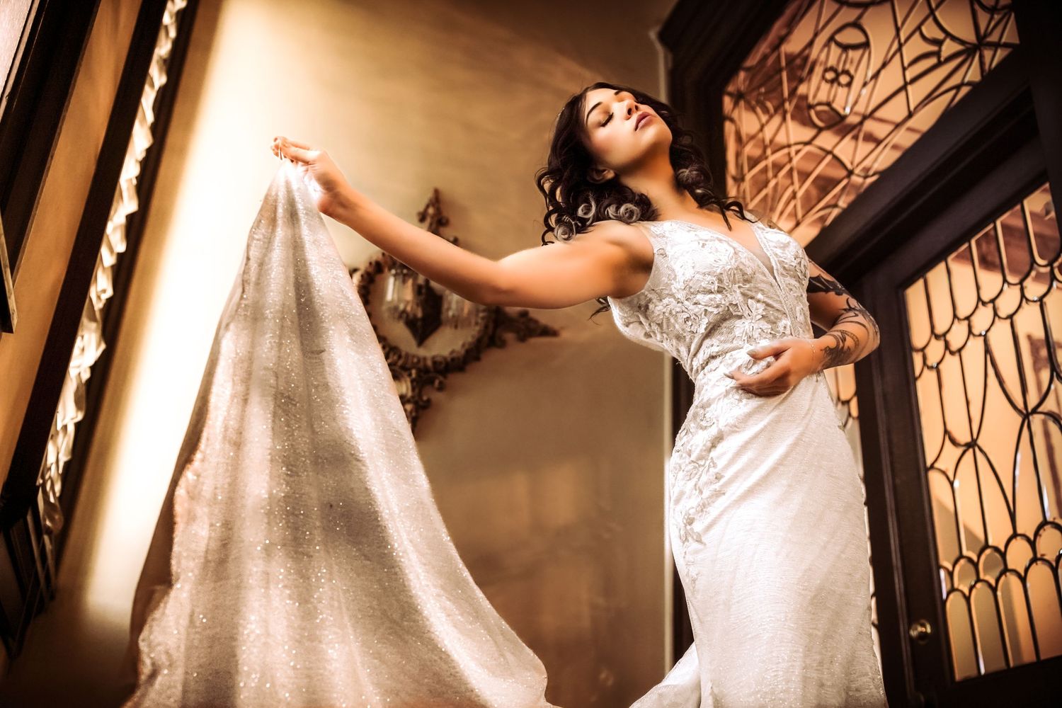 A bride in an elegant white lace wedding dress poses dramatically while holding flowing fabric in a warmly lit hallway.
