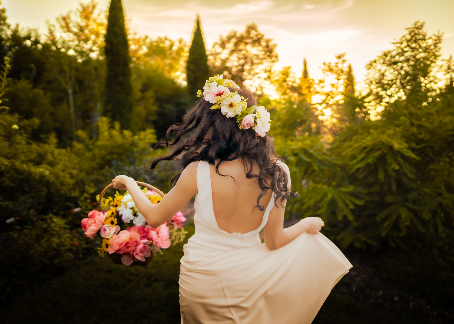 White-dressed figure twirls with flowers in garden at sunset among cypress trees.