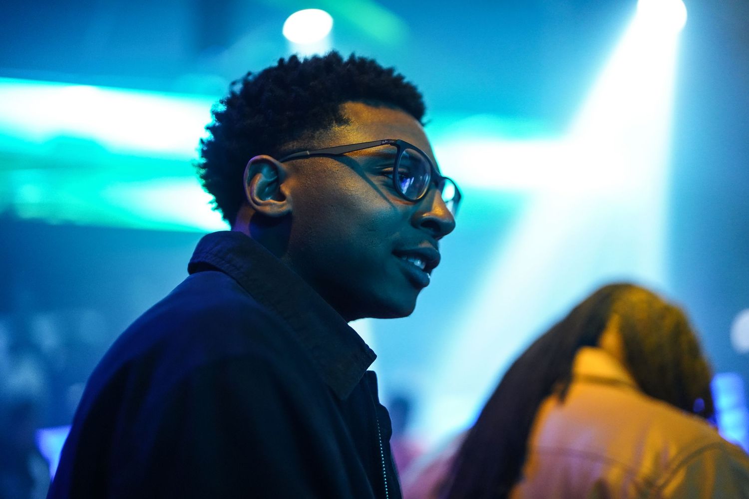 A performer with glasses and curly hair stands in profile against blue stage lighting at a concert venue.